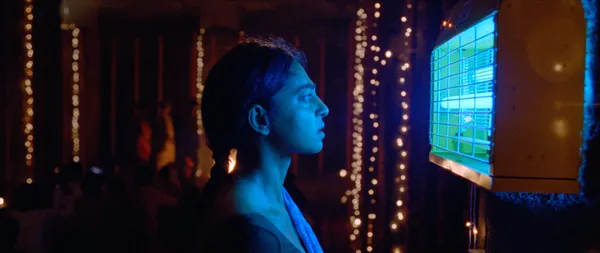 A still from the film Sister Midnight. A young Indian woman stands in an outdoor space illuminated by vertical strings of fairy lights. it is night time and she is staring into a glowing fan.