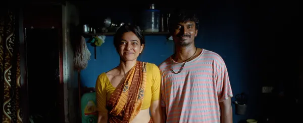 A still from the film Sister Midnight. A smiling Indian wife and husband stand side by side in a blue-painted kitchen.