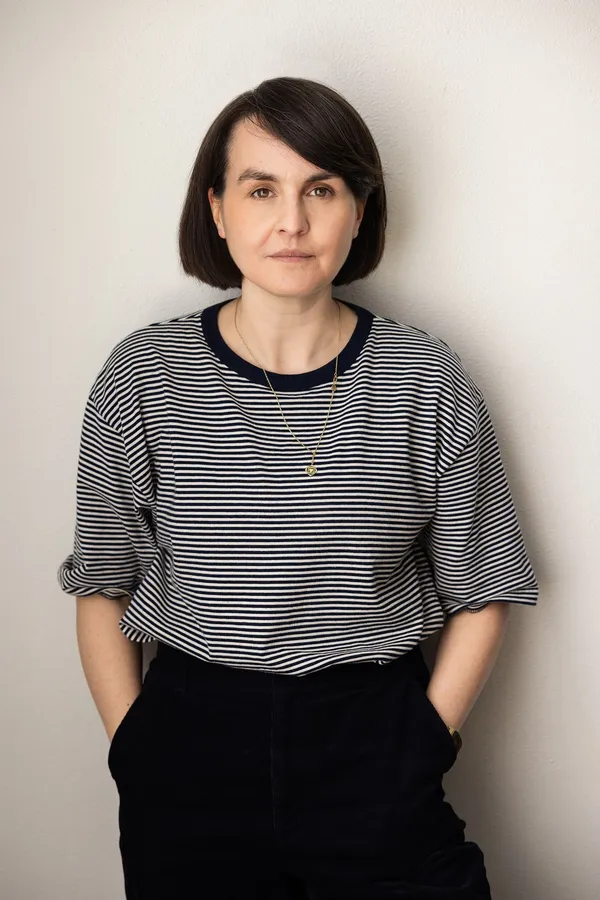 A woman with light coloured skin and dark hair, dressed in black trousers and a top with narrow black and white stripes.