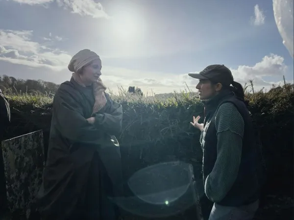 A tall white woman in a big coat, with a wrap around her hair, stands in a ditch taking instruction from a smaller US-Iranian woman.