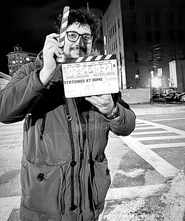 A behind the scenes shot from the film Stationed At Home. On a snowy night, a smiling white man holds up a clapperboard.