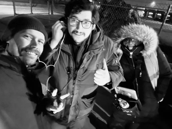 A behind the scenes shot from the film Stationed At Home. Three white men in warm winter clothing huddle together. Two of them are sharing earphones.