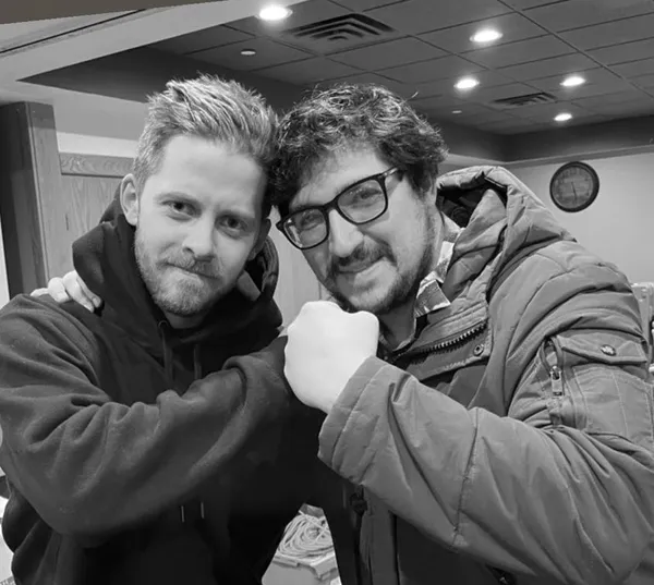 A behind the scenes shot from the film Stationed At Home. Indoors, two white men in warm jackets bump fists.