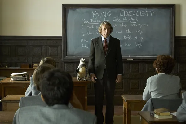A still from the film The Penguin Lessons. In a classroom, boys in grey uniforms watch a middle-aged white teacher stand in front of a blackboard with the words 'young idealistic' above the poem Sea Fever by John Masefield, beside a penguin.