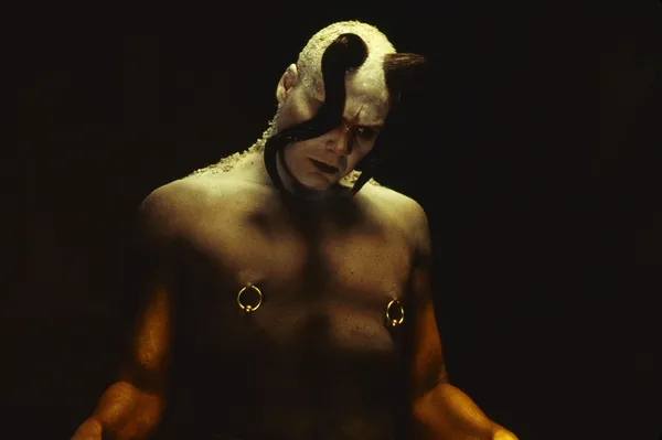 A still from the film The Cell. A masculine figure with a naked torso, nipple rings, and a horn and tentacles emerging from its head.
