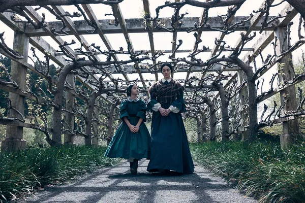 A still from the film Mārama. A Māori woman and girl walk through an arbour in spring, whenm buds are beginning to form on twisted branches.