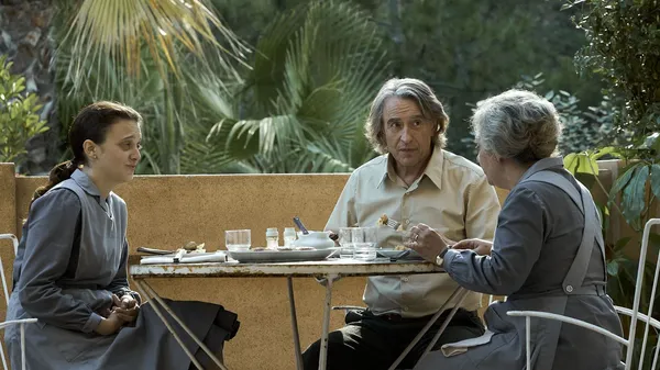 A still from the film The Penguin Lessons. Two Argentinean women, one young and one old, wearing grey cleaners' uniforms, share a light meal with a middle aged white man on a small balcony behind which thick foliage grows.
