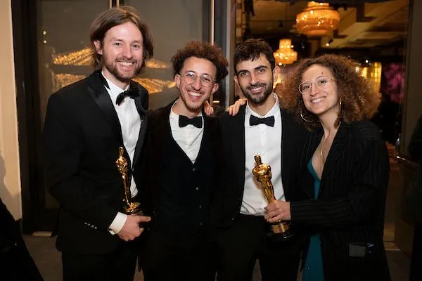 Three men and a woman stand in a lobby area, wearing evening dress, with broad grins on their faces. Two of them are holding Oscar statuettes.