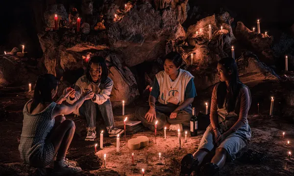 A still from the film Amoeba. Four casually dressed Singaporean teenage girls sit around in a cave full of candles, with a bottle of wine.