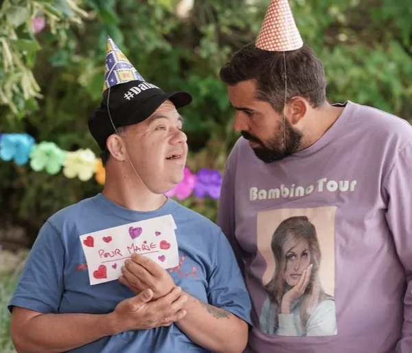 A still from the film A Little Something Extra. Two white men wearing pastel-coloured t-shirts and party hats stand in a garden which has been decorated with bunting.  The shorter one, who looks as if he has Down syndrome, is holding a letter.