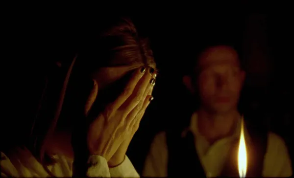 A still from the film The Assistant. A blond white woman with dark nail polish stands beside a candle, covering her face with her hands; a white man in shirt and waistcoat can dimly be seen beyond the flame.