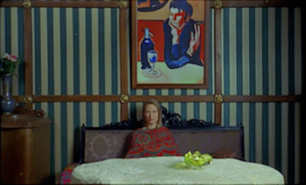 Agnieszka Zulewska. A middle-aged white woman sits in a large bed low down in the frame in a room with blue and white vertically striped wallpaper.