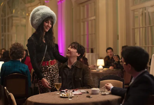 A still from the film The Wizard Of The Kremlin. In a crowded restaurant, a young man sits with his arm around a young woman in leopard print leggings and an enormous fur hat, who is standing.