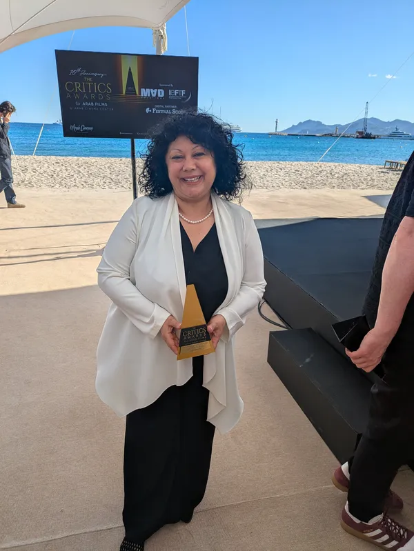 The film critic Alin Taşçıyan. A Turkish woman stands on a sunny terrace by the sea, smiling broadly as she holds an award.