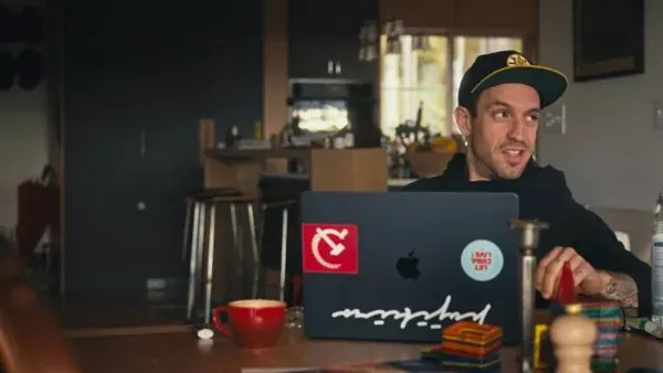 A still from the film All About The Money. In a kitchen, a young white man works at a black laptop with a bright red hammer and sickle sticker on it.