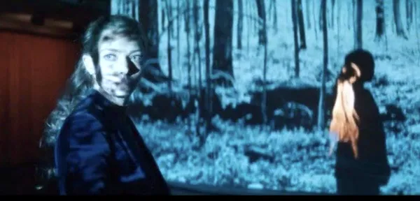 A still from the film Seven Veils. Figures stand in woodland on a projected, blue-tinted screen.