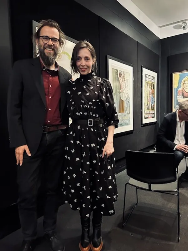 A smiling, bearded, bespectacled white man stands beside a smiling white woman inb a dark-walled room lined with framed posters.