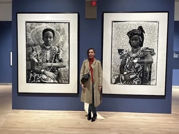 A middle-aged white woman in a long beige coat stands in an art gallery between two large photographic prints of African women.