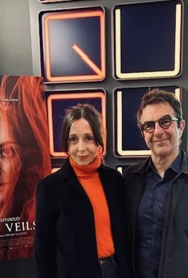 A middle aged white woman wearing an orange jumper and a dark jacket stands beside a middle aged white man in a dark suit. behind them, in a square shape, are the letters Q U A D.