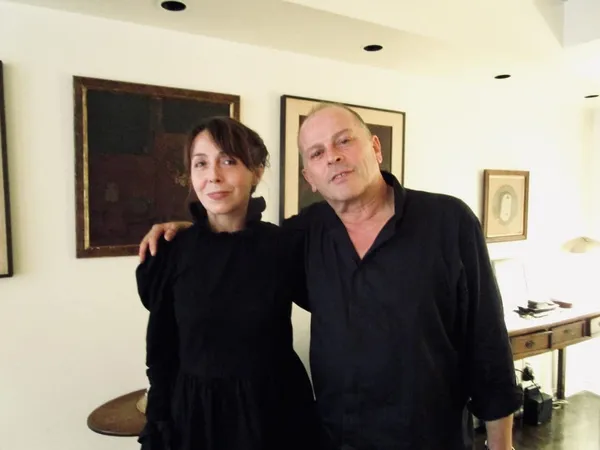 A middle aged white woman and man, both wearing black, stand in a long white room with framed paintings on the walls.