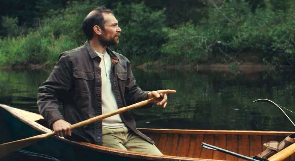 A still from the film Who By Fire. A middle aged white man rows a boat along a river whose bank is thick with trees.