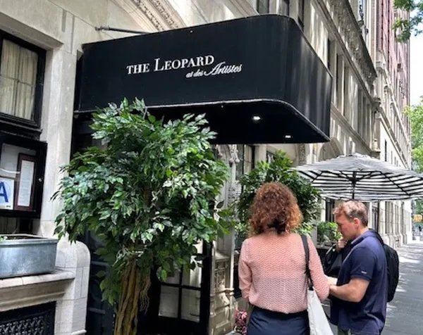 The entrance to a restaurant on a terrace street. A slender potted tree stands beside a sign which says 'The Leopard at Des Artistes'.