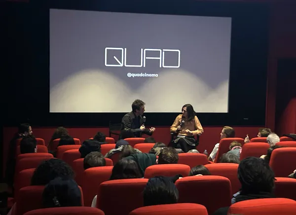 A white man and a white woman sit in front of a cinema screen which bears the word 'quad'. A small audience of people in red velvet cinema seats is watching them.