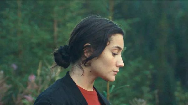 A still from the film Who By Fire. The head and shoulders of a light-skinned young woman with black hair in a bun.