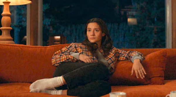 A still from the film Who By Fire. A young woman sits on an orange sofa in front of a window at night.