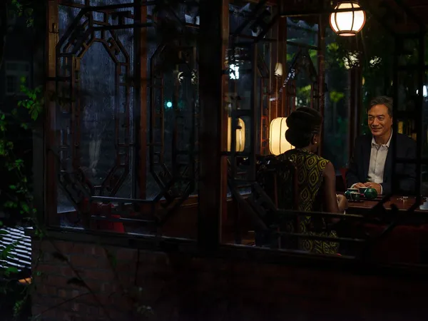 A still from the film Black Tea. One corner of a tea shop is seen from outside at night. A Black woman can be seen from behind as she sits at a small table with an East Asian man.