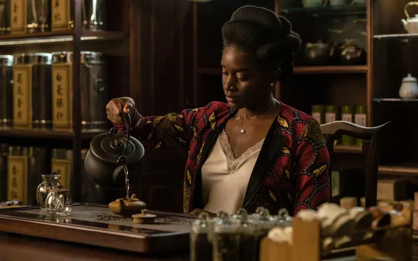 A still from the film Black Tea. A young Black woman stands behind the counter of a tea shop, carefully pouring tea from a kettle into a small pot.