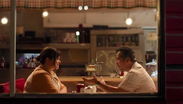 A still from the film Bob Trevino Likes It. A young white woman and a middle aged white man laugh together in a warmly lit café.