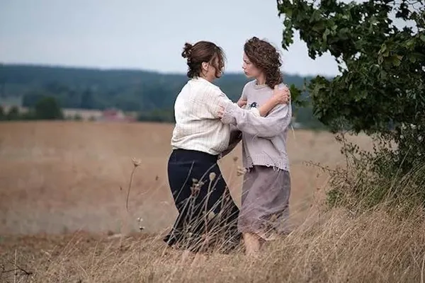 A still from the film Mirrors No. 3. In a field with a forest beyond it, a middle-aged white woman and a younger white woman grapple with one another in what could be a fight and could be a hug.