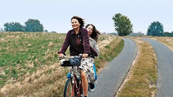 A still from the film Mirrors No. 3. A white woman cycles along a country road with a white girl on the back of her bicycle.