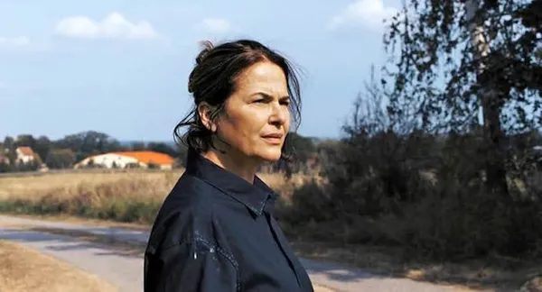 A still from the film Mirrors No. 3. A dark haired white woman stands by the wide of a country road, squinting into the distance.