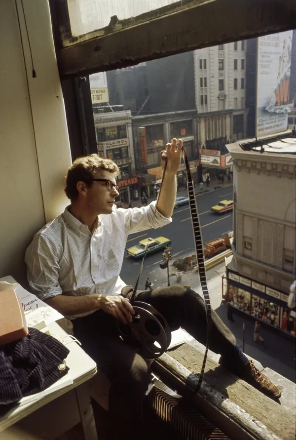 A still from the film Being Bo Widerberg. A thin, bespectacled white man sits in a sunny window above an urban street, holding a roll of film and stretching it out to peer at individual frames.