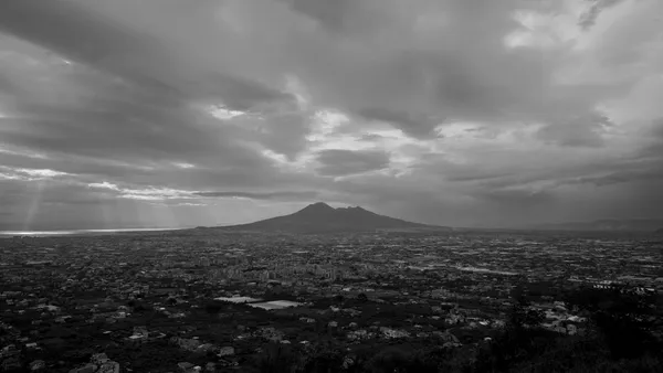 A black and white still from the film Below The Clouds. A city lies spread out on a wide plain around a volcano.