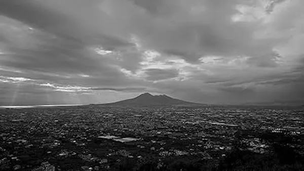 A black and white still from the film Below The Clouds. A city lies spread out on a wide plain around a volcano.