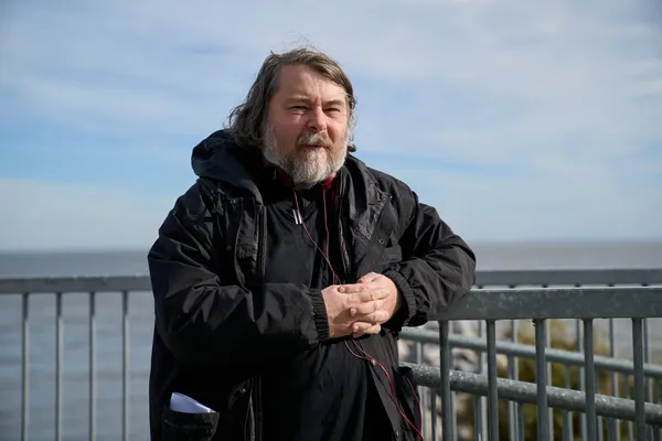 The filmmaker Ben Wheatley. A middle-aged white man with black clothes, shoulder-length hair and a greying beard stands beside railings, with a calm sea beyond them.