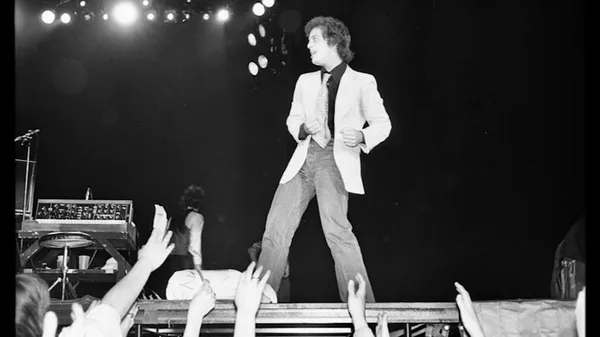 A black and white image of the musician Billy Joel performing onstage, wearing a white jacket, with fans stretching their arms up towards him.