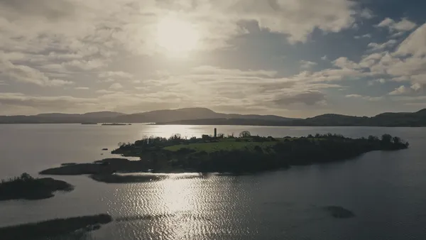 Inis Cealtra, a low grassy island with scattered trees and a tall stone column, situated amid the placid silvery waters of a wide lough, under a low sun.