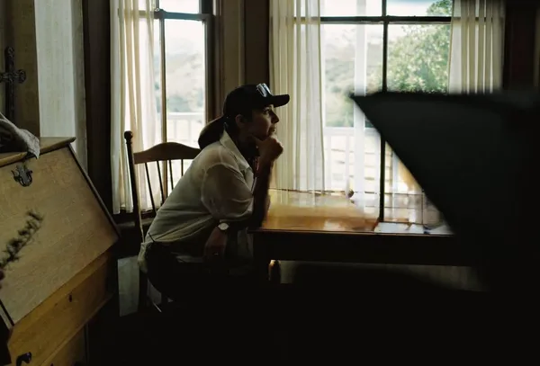 A US-Iranian woman sits at a table in the bay window of a room crowded with furniture. She is wearing a peaked cap, has a radio in her back pocket and looks keenly attentive.