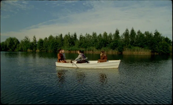 A still from the film The Assistant. Four white people sit in a white rowing boat, out on a conifer-lined lake on a sunny day.
