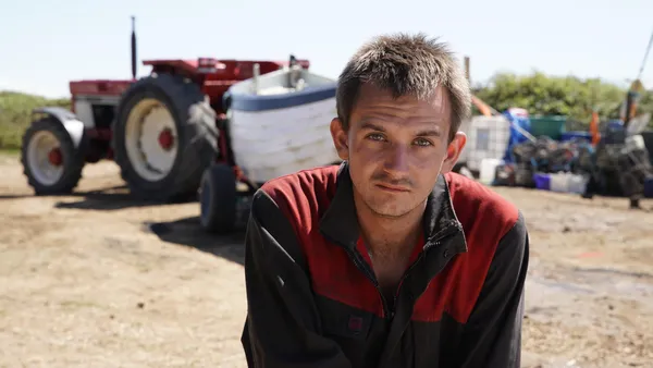 Brandon Vlieghe. A man in a red and black top stands in a yard. A tractor towing a boat is parked in the background.