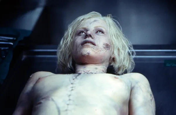 A still from the film The Cell. A white androgynous figure with bleached hair lies on a metal gurney.