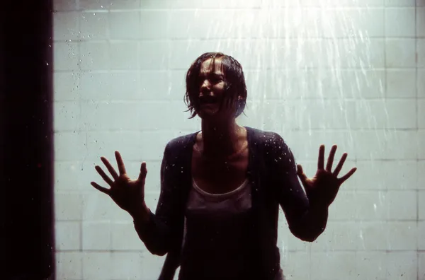 A still from the film The Cell. A dark-haired white woman wearing a white t-shirt and dark cardigan takes a shower, pressing her palms against the glass in front of her.