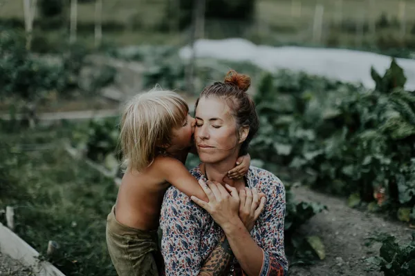 A still from the film A New Kind Of Wilderness. A white woman with tattoos along one forearm, wearing a floral dress, kneels in a vegetable garden, her eyes closed as a white child hugs her tightly and kisses her.