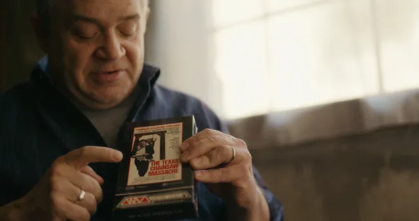 Patton Oswalt, a smiling, grey-haired middle aged man, holds up a VHS copy of Texas Chain Saw Massacre.