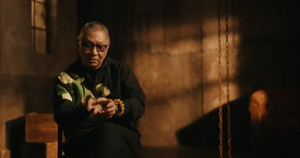 Takashi Miike, a bespectacled older Japanese man, sits in a room with chains hanging down beside him.