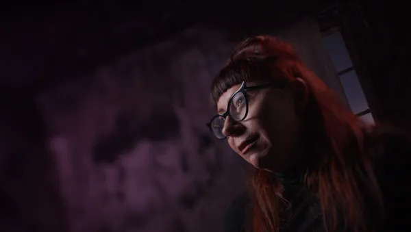 Alexandra Heller-Nicholas, a bespectacled white woman with dark crimson hair, sits in a purple-lit room.